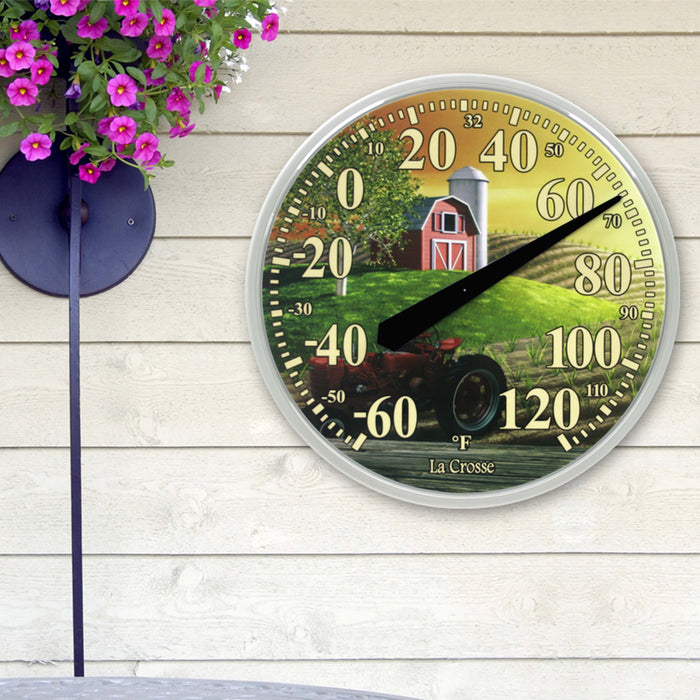 Lifestyle image of the 104-114-FARM 13.5-inch Dial Thermometer - Barn, with key hider, temperature scale in Fahrenheit -60 to 120 degrees with a red-tipped pointer in a white slimline case hanging on a white wood siding house next to pink flowers