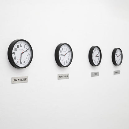 Lifestyle image of the 404-3020B-4TZ Four Pack 8-inch Basic Wall Clock with City Plaques. Four black 8-inch round wall clocks with white faces and black numerals are mounted in a horizontal row on a light gray wall. Each clock has a red second hand and displays a different time. Beneath each clock is a small rectangular plaque labeling a city: “Los Angeles,” “New York,” “Paris,” and “Tokyo,” indicating different time zones.