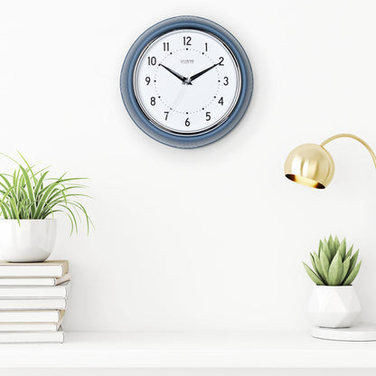 Lifestyle image of the 404-3024IB 9.5-inch Diner Clock – Indigo Blue shown hanging on a white wall above a white shelf with plants, books, and a retro lamp. The clock features a retro-style Indigo Blue frame with a white dial, chrome bezel, black numbers and minute markings, black metal hands, and a silver second hand.