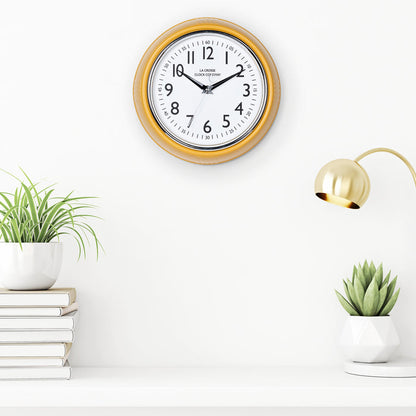 Lifestyle image of the 404-3024MY 9.5-inch Diner Clock – Mustard Yellow shown hanging on a white wall above a white shelf with plants, books, and a retro lamp. The clock features a retro-style mustard yellow frame with a white dial, chrome bezel, black numbers and minute markings, black metal hands, and a silver second hand.