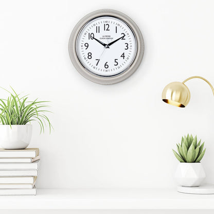 Lifestyle image of the 404-3024TP 9.5-inch Diner Clock – Taupe shown hanging on a white wall above a white shelf with plants, books, and a retro lamp. The clock features a retro-style taupe frame with a white dial, chrome bezel, black numbers and minute markings, black metal hands, and a silver second hand.
