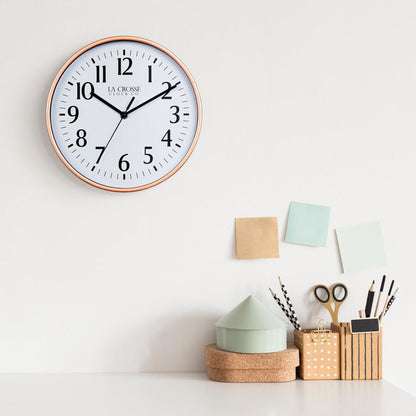 Lifestyle image of the 404-3025C 10-inch Basic Wall Clock - Copper hanging on a white wall above a white desk with cork, wood, and pastel desk accessories. The clock features a white dial, black numbers and markings, and hour, minute, and second hands, surrounded by a copper finish flush-mount frame.
