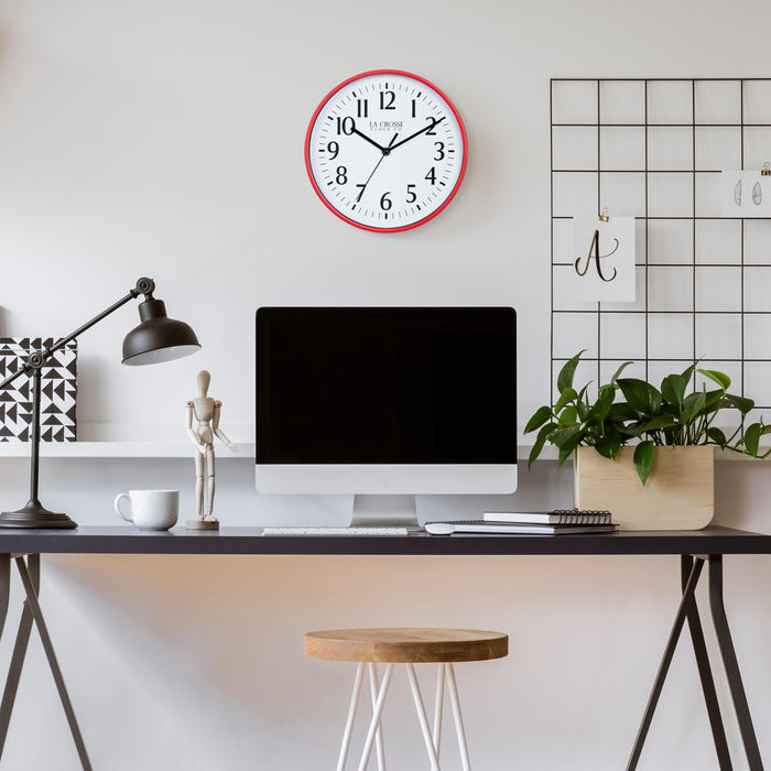 Lifestyle image of the 404-3025R 10-inch Basic Wall Clock – Red, hanging on a light tan wall above a black desk. The desk has a computer, notebooks, a plant, a coffee mug, a black desk lamp, and a posable wood model. The clock features a white dial, black numbers, markings, hour, minute, and second hands surrounded by a red flush-mount frame.