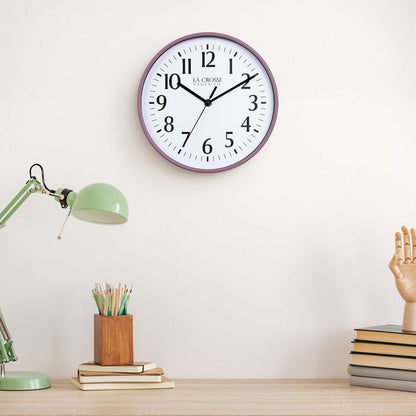 Lifestyle image of the 404-3025RR 10-inch Basic Wall Clock – Ruby Red, hanging on a light tan wall above a light wood desk with a green desk lamp, a wood pencil holder on top of a stack of notebooks, and a wood modeling hand sitting on top of a pile of books. The clock features a white dial, black numbers, markings, hour, minute, and second hands surrounded by a ruby red rose flush-mount frame.
