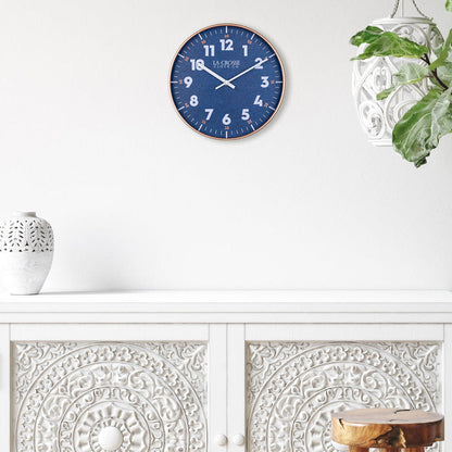 Lifestyle image of the 404-3027C Portside eleven inch navy blue wall clock with white numbers, orange time markings, and a copper case featuring white hour and minute hands over a buffet side table of white with a carved white planter and vase
