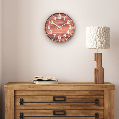 Lifestyle image of the 10.8-inch Atwood Wall Clock hanging above a rustic wood dresser with a wood lamp. The clock features a copper finish frame, a wood design dial, white numbers, and white metal hour and minute hands.