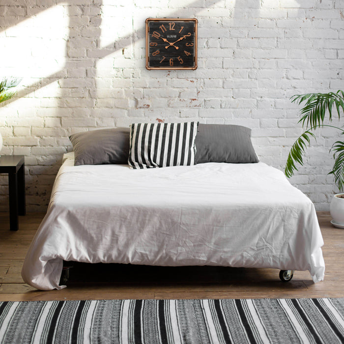 Lifestyle image of the 404-3412D 16-inch Foundry Metal & Wood Wall Clock - Dark, shown hanging on a white-washed brick wall above a bed. The clock features a square metal pipe frame in a copper finish, a black-stained wood slat clock face with copper-finished metal numbers and hands.