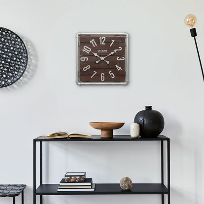 Lifestyle image of the 404-3412L 16-inch Foundry Metal & Wood Wall Clock - Light, shown hanging above a black metal shelving unit with books and vases. The clock features a square metal pipe frame in a weathered pewter finish, a truffle-brown-stained wood slat clock face with pewter-finished metal numbers and hands.