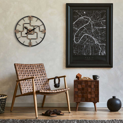Lifestyle image of the 404-3420 20-inch Pipeworks Metal Wall Clock shown hanging on a tan plaster finish wall hanging next to map artwork, and above a dark wicker chair with a woven wood side table. The clock features an open face dial with a weathered bronze and antique brass finish pipe framework, and antique brass numbers 3, 6, 9, and 12.