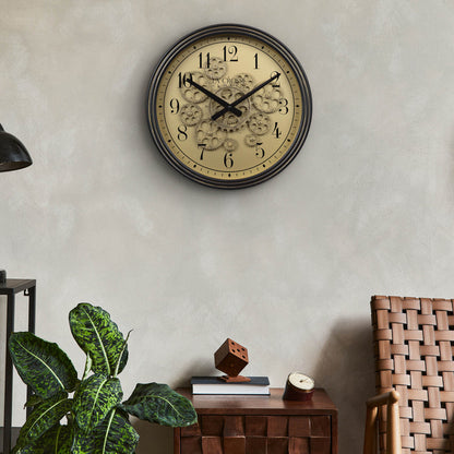 Lifestyle image of the 404-3439 15-inch Moving Gears Wall Clock hanging on a wall above a chair and side table with a tropical theme decor. The clock features a metal oil-rubbed antique frame, distressed brushed dial with moving gears, and black numbers and hands.