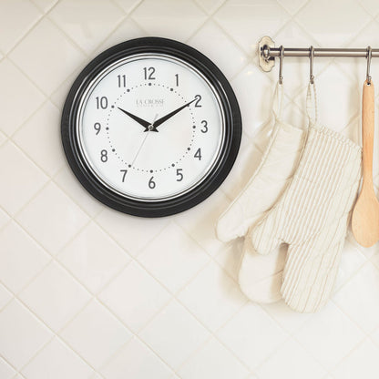 Lifestyle image of the 404-3624B 9.5-inch Diner Wall Clock hanging on a white tile wall in a kitchen. The clock features a black frame with a chrome bezel accent, an off-white dial with black numbers and hour dots, a silver second hand, and black hour and minute hands.