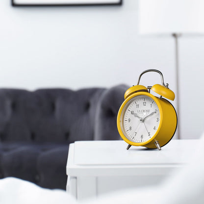 Lifestyle image of the 617-3314MY Metal Twin Bell Alarm Clock – Mustard Yellow, sitting on a white side table with a dark grey couch in the background. The clock features a white dial with black numbers, silver hands and accents, in a mustard yellow metal case.