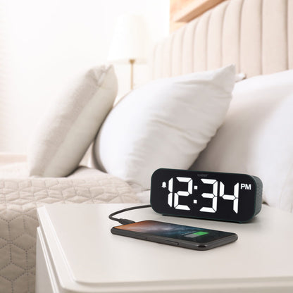Lifestyle image of the 617-714G Insta-Set Digital LED Alarm Clock with Bluetooth® Technology - Green sitting on a white nightstand with a bed in the background. The alarm clock is shown with a smartphone being charged. The alarm clock features large white digits, a PM indicator, and an alarm icon in a green case.