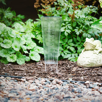 704-5331 USA-Made Clear Measure Rain Gauge lifestyle in a plant bed with bark colored rocks green foiliage and a frog sculpture