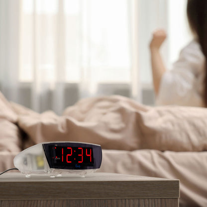 Lifestyle image of the 75908 0.9-inch Red LED Clear Case Alarm Clock shown on a night table next to a bed. The digital alarm clock features red digits with a black lens, PM and ALARM indicators, and is in a clear case.
