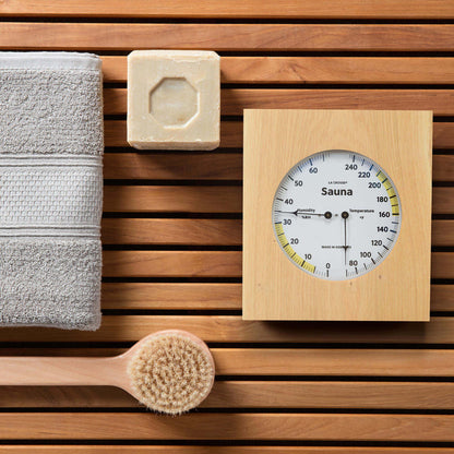 Lifestyle image of the 914-620 Sauna Thermometer and Hygrometer dial in oiled oak wood casing displaying a dial split in half with temperature on the right in Fahrenheit from 60 degrees to 245 degrees with optimal range in yellow and the left half showing humidity readings from 0 percent to 75 percent with the optimal range highlighted in yellow sitting on a sauna bench with a towel brush and soap