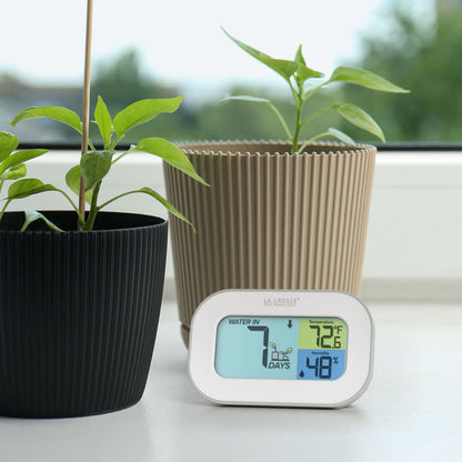 Image of the 919-147 Digital plant care timer with temperature and humidity. The timer sits on a windowsill with potted plants. Battery-operated, the timer features a white and gray case with a three section screen. The turquoise blue section displays how many days since the last watering (plant graphic with an up arrow) the yellow section lists the temperature at 72.6° F, and the dark blue section lists the humidity level at 48 percent with a water drop icon.